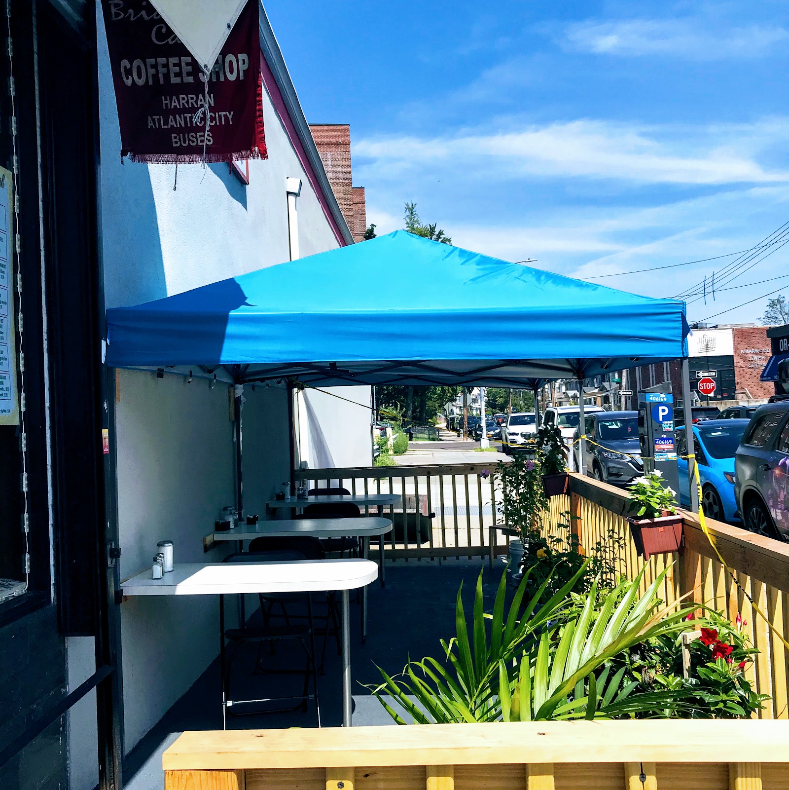 The sidewalk deck beside the cafe: four tables under a bright blue canopy, a new timber rail with planters of flowers, and Queens Boulevard traffic beyond. A second awning above reads Coffee Shop, Harran, Atlantic City, Buses