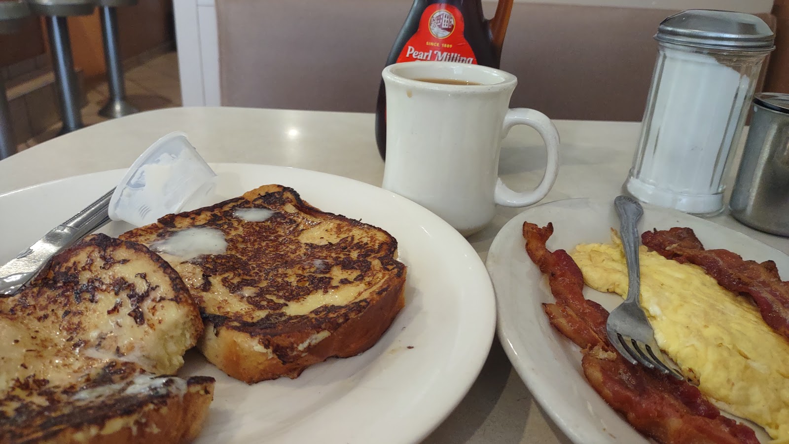 Two thick slices of griddled French toast on a white plate, a mug of black coffee and a bottle of syrup behind, and a second plate of bacon and folded omelette to the side, with a glass sugar shaker on the table