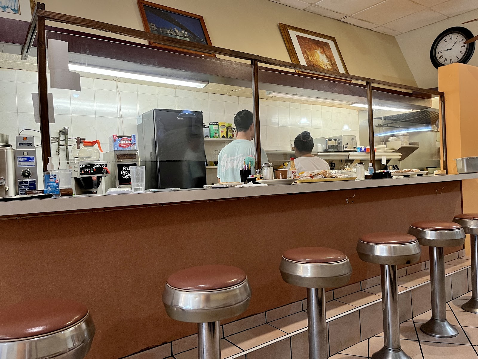 The counter inside: five chrome-pedestal stools with brown vinyl seats in a row, a long formica counter, and behind it a pass-through window into the kitchen where two cooks are working under strip lights, with coffee brewers and a wall clock to one side