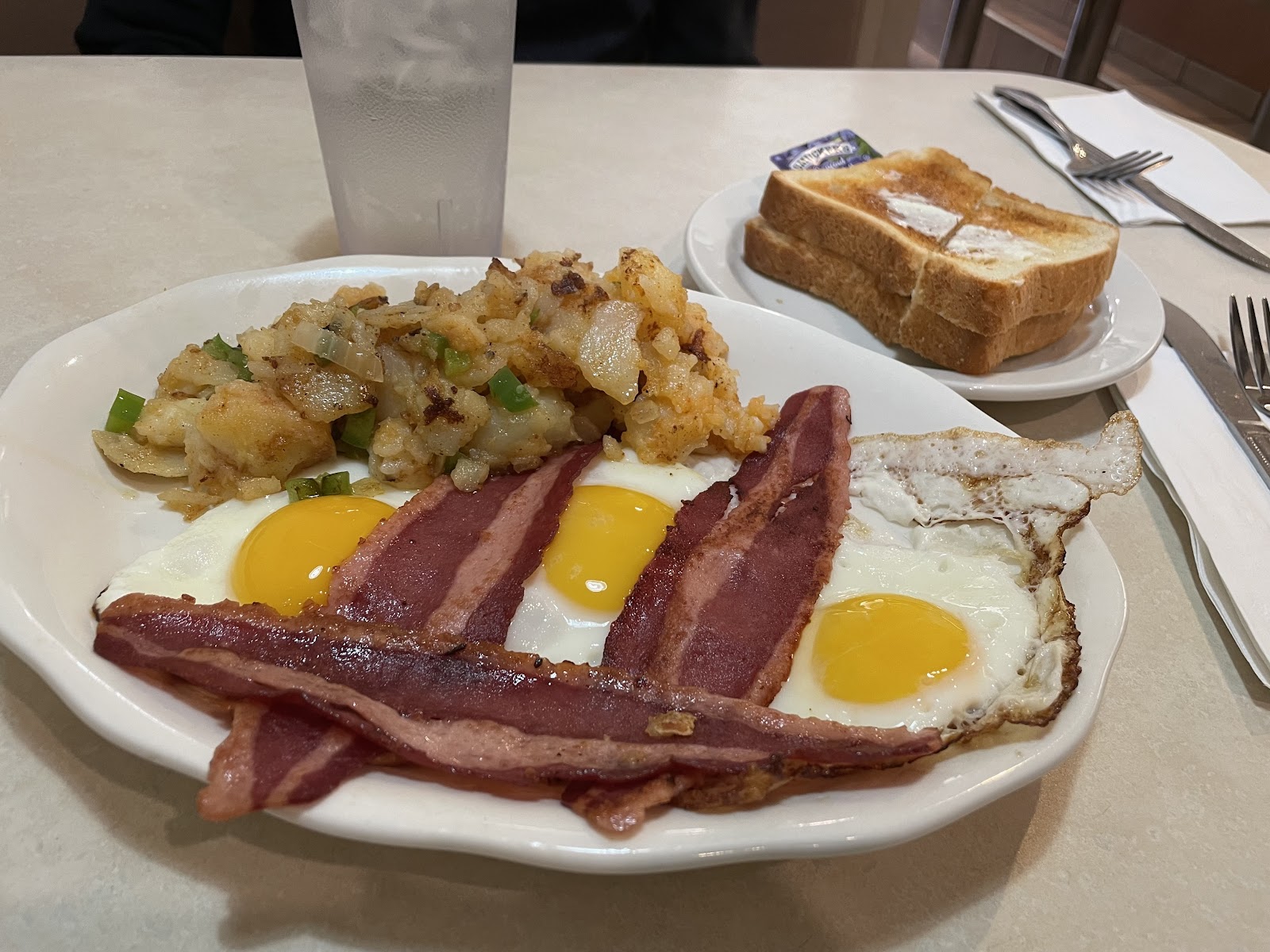 Two eggs sunny side up with three rashers of bacon laid across them and a pile of home fries flecked with green pepper and onion, on a white oval plate, with buttered white toast on a side plate