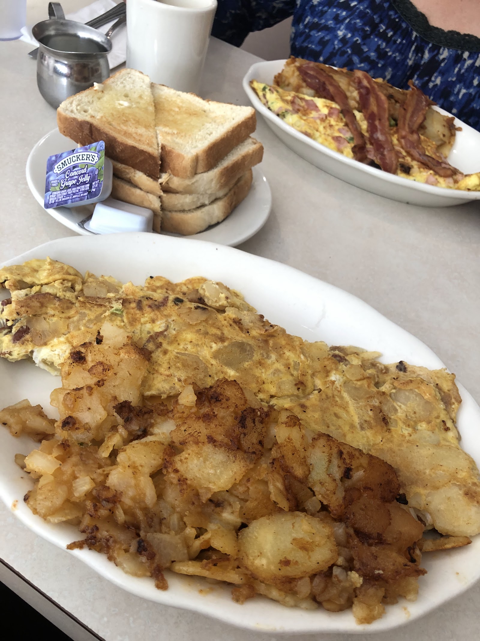 A large folded omelette almost buried under home fries on a white oval plate, with a stack of toast and a packet of grape jelly on a side plate and a second omelette with bacon beyond