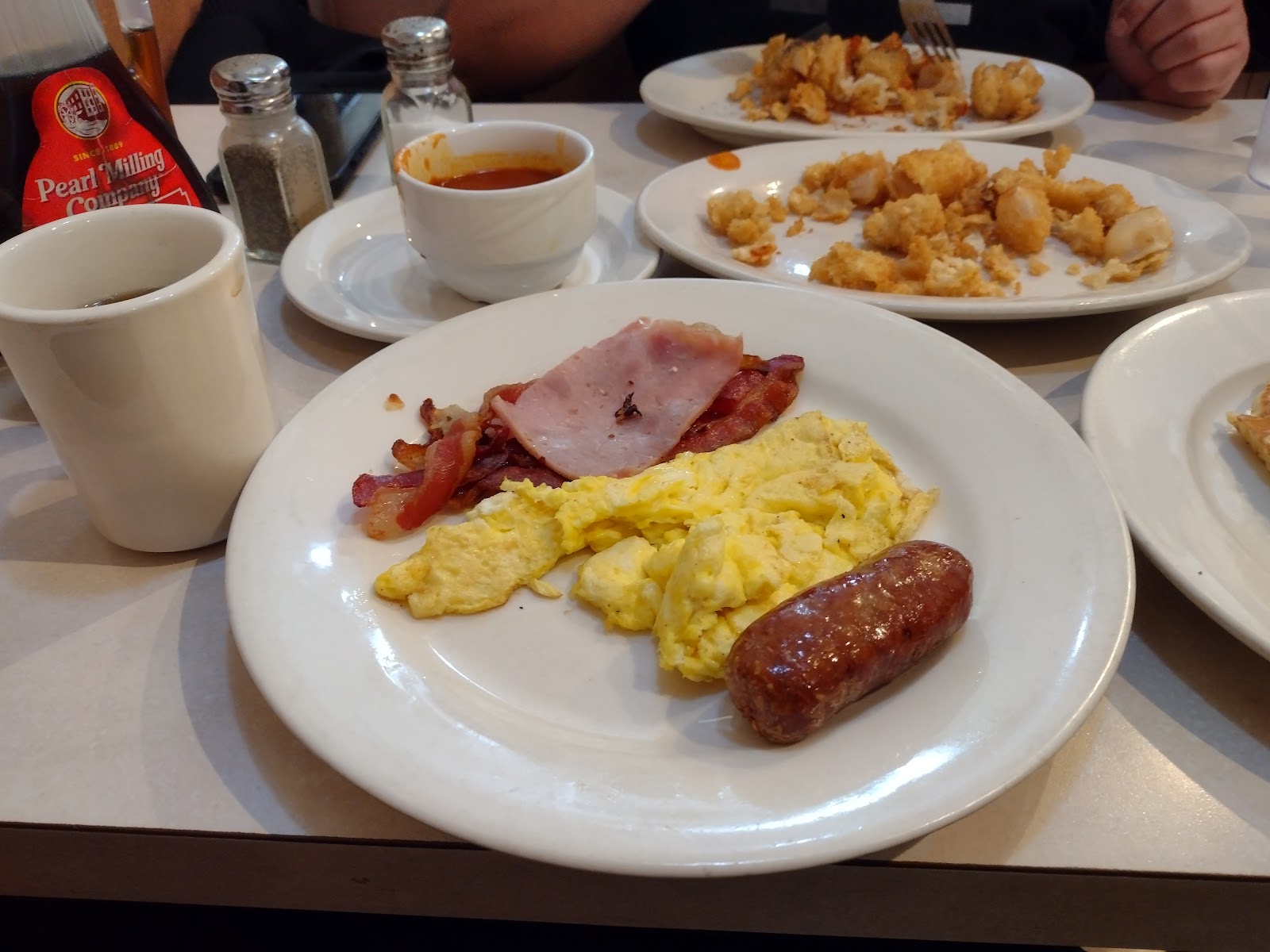 Scrambled eggs with grilled ham, bacon and a sausage link on a white plate, with a cup of soup, a mug of coffee and two shared plates of golden fried seafood on the table behind