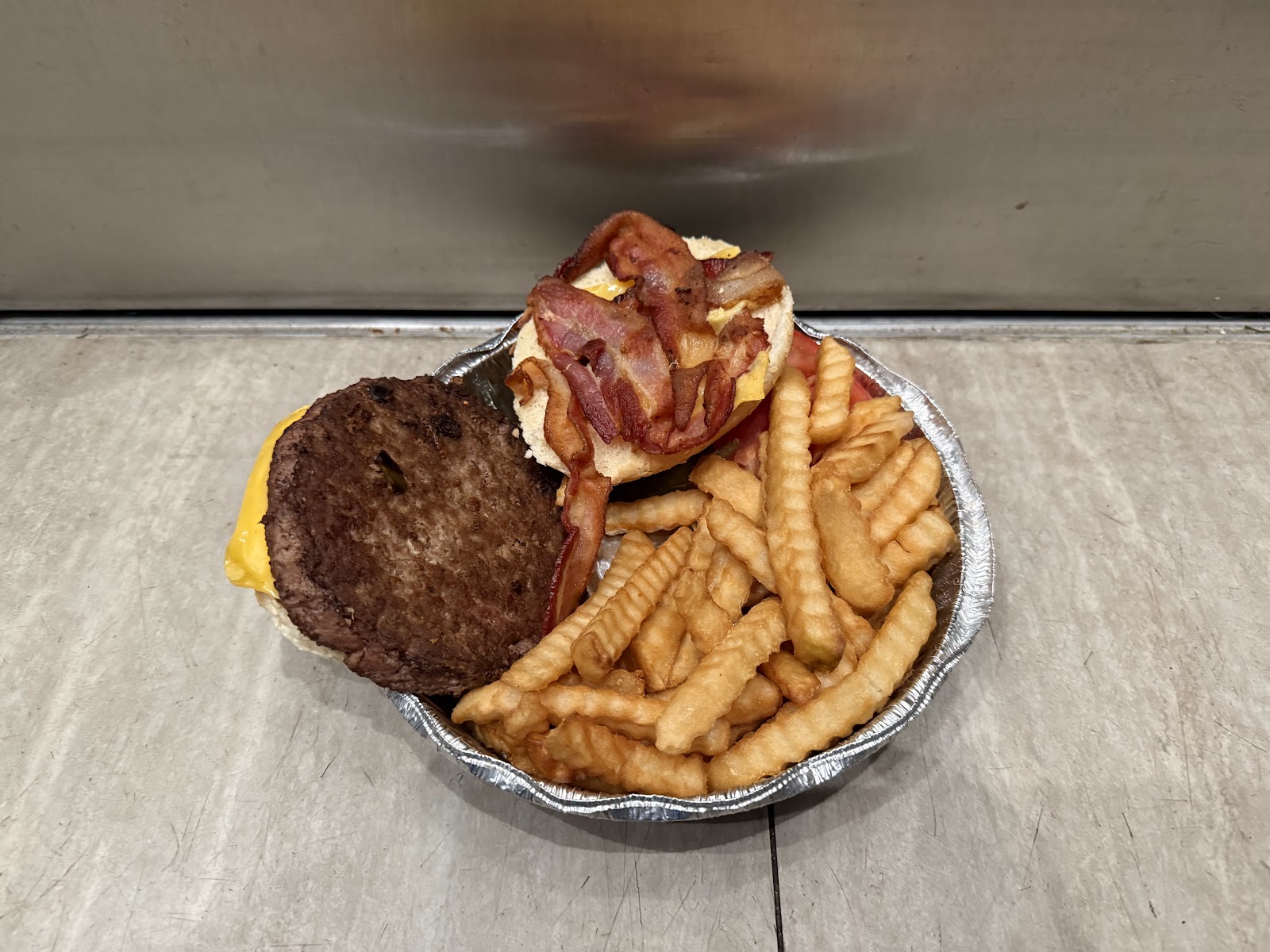 A takeout bacon cheeseburger in a foil container, the patty and melted cheese laid open beside the bacon-topped bun half, with crinkle-cut French fries filling the rest of the dish